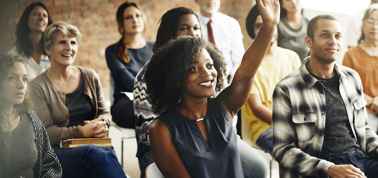 An audience of casually dressed people looking towards a speaker, with a female raising her hand.